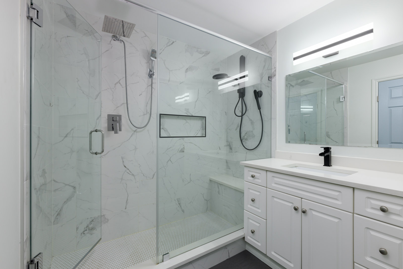 Modern bathroom with a glass-enclosed shower, marble-patterned walls, dual showerheads, a white vanity with cabinets, and a rectangular mirror with overhead lighting.